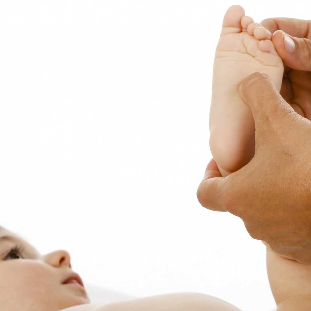 Ein Baby liegt auf dem Rücken und schaut zu, wie es am linken Fuß behandelt wird von der Osteopathin